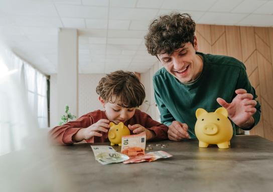 Vater und Sohn sitzen lächelnd am Tisch auf dem Geld und 2 Sparschweine liegen.
