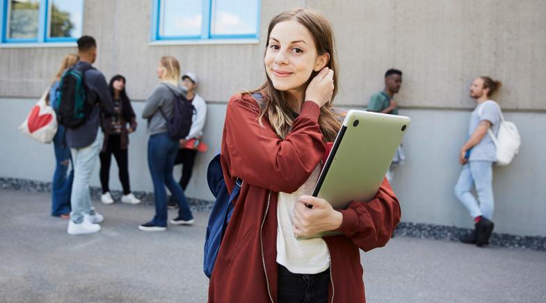 Studentin steht vorm Universitätsgebäude mit Laptop und lächelt