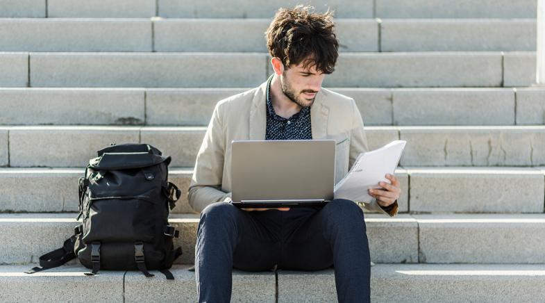 Student sitzt auf einer Treppe mit Laptop auf dem Schoß und schaut auf einen Spiralblock in seiner Hand.