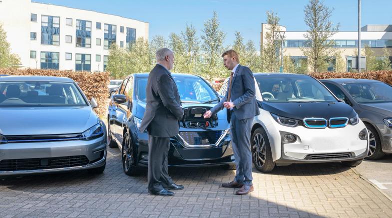 Autoverkäufer zeigt einem Kunden auf dem Parkplatz wo man ein E-Auto auflädt.