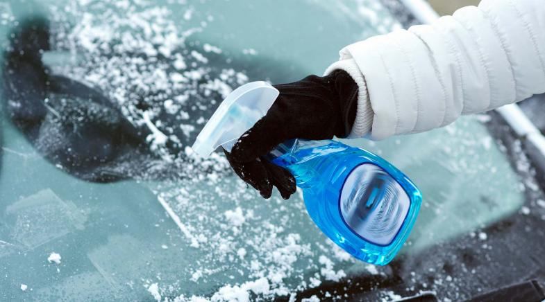 Eine Person sprüht mit einer blauen Flasche Enteisungsmittel auf eine vereiste Windschutzscheibe.