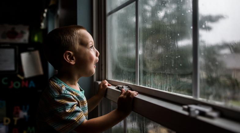 Ein Kind schaut fröhlich durchs Zimmerfenster auf den Regen draußen.