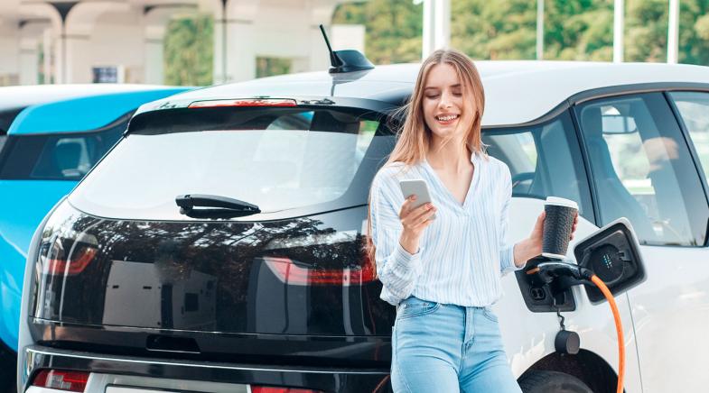 Frau schaut auf ihr Handy und hält einen Kaffeebecher in der hand während ihr Elektroauto neben Ihr an einer Ladesäule lädt.