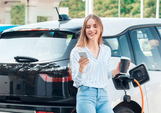 Frau schaut auf ihr Handy und hält einen Kaffeebecher in der Hand während ihr Elektroauto neben Ihr an einer Ladesäule lädt.
