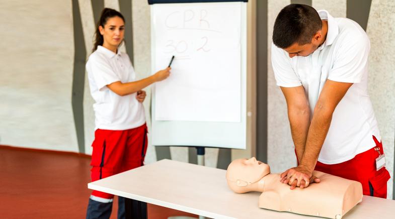 Eine Sanitäterin zeigt auf ein Flipchart und ein Sanitäter macht eine Herzdruckmassage an einer Erste-Hilfe-Puppe.