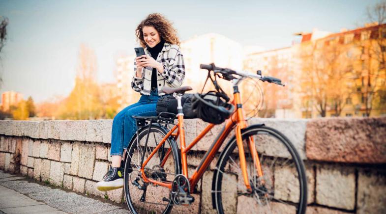 Frau sitzt auf einer Mauer und schaut auf Ihr Handy. Neben Ihr lehnt ihr Fahrrad an der Mauer.