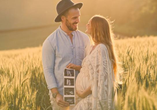 Ein Paar steht in einem Weizenfeld und hält Ultraschallbilder ihres Babys in der Hand.