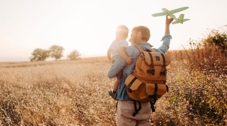 Ein Mann steht auf einer Wiese mit einem Baby im Arm und einem Modellflugzeug in einer Hand.