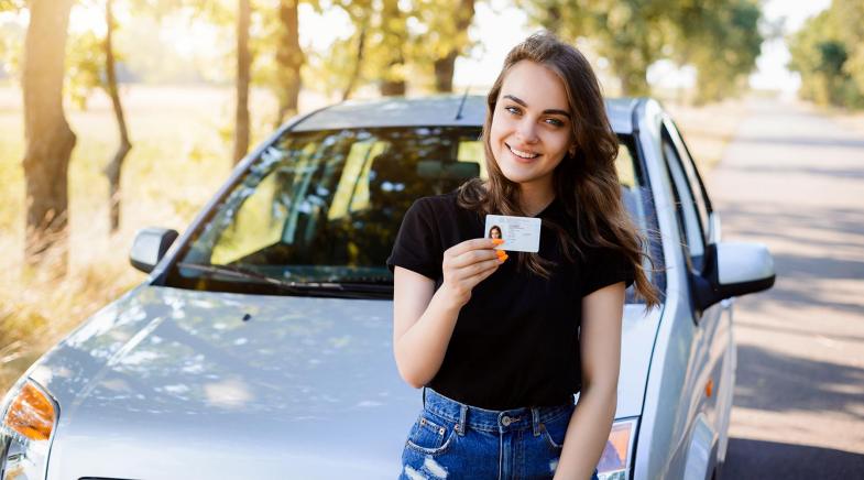 Junge Frau lehnt an ihrem Auto, das am Straßenrand einer Allee parkt, und zeigt ihren Führerschein.