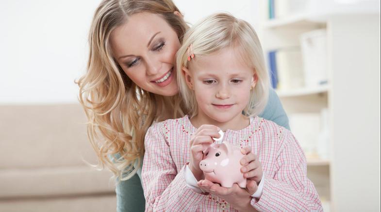 Eine Mutter sitzt hinter ihrer Tochter und schaut ihr dabei zu wie sie Geld in ein Sparschwein wirft.