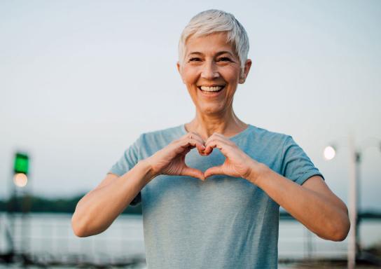 Eine Frau am Meer formt mit ihren Händen ein Herz und lacht.