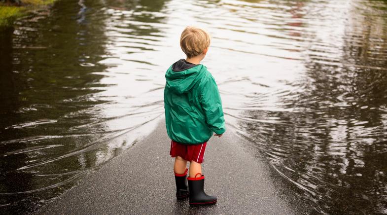 Kleiner Junge mit Regenjacke und Gummistiefeln steht auf einer überschwemmten Straße.