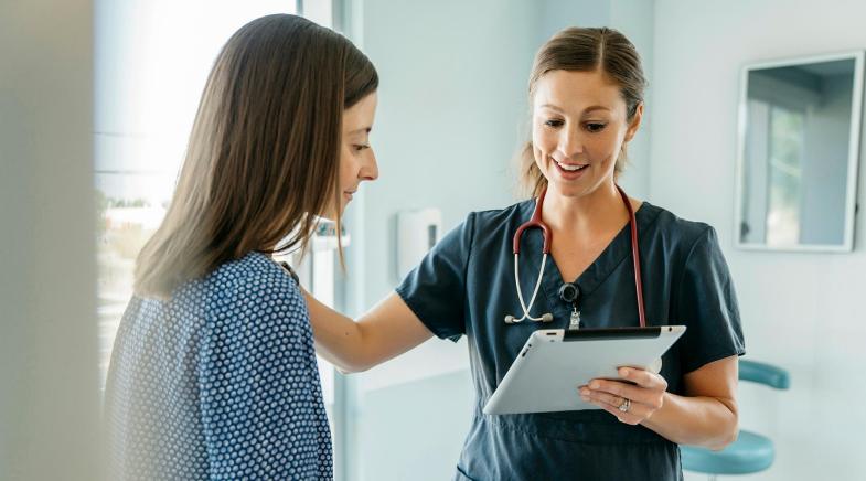 Ärztin schaut im Untersuchungszimmer auf ein Tablet in ihrer Hand und redet lächelnd mit einer Patientin.