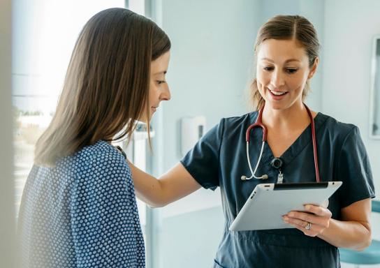 Ärztin schaut im Untersuchungszimmer auf ein Tablet in ihrer Hand und redet lächelnd mit einer Patientin.