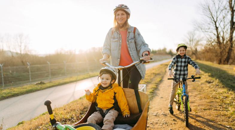 Eine Mutter mit zwei Kindern fährt Lastenfahrrad. Ein Kind fährt mit einem Kinderrad nebenher, das zweite sitzt im Lastenrad.