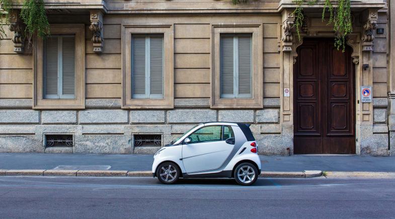 Ein weißes Microcar parkt seitlich in der Stadt am Straßenrand vor einem Haus.