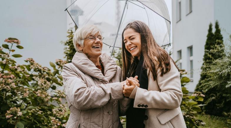 Eine alte und eine junge Frau stehen unter einem Regenschirm und lachen.