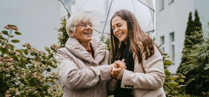 Eine alte und eine junge Frau stehen unter einem Regenschirm und lachen.
