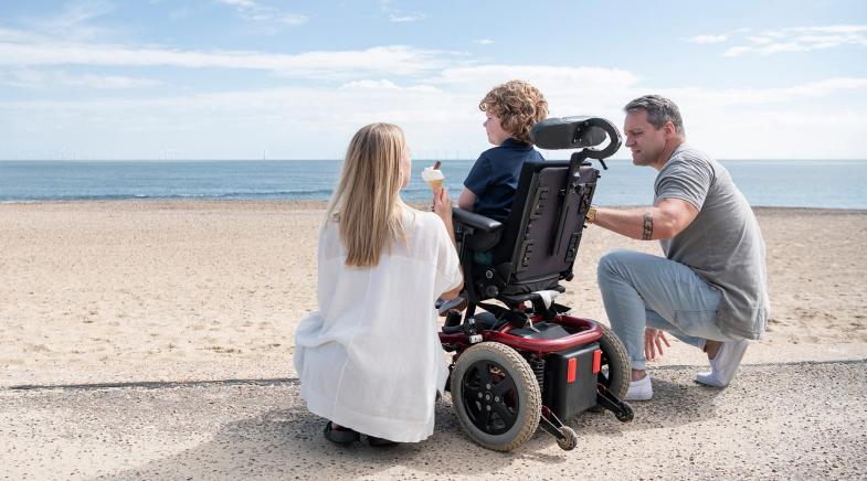 Elternpaar mit einem Kind im Elektrorollstuhl am Strand.