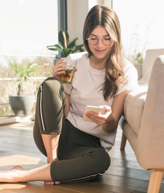 Junge Frau sitzt entspannt vor ihrem Sofa und schaut aufs Handy.