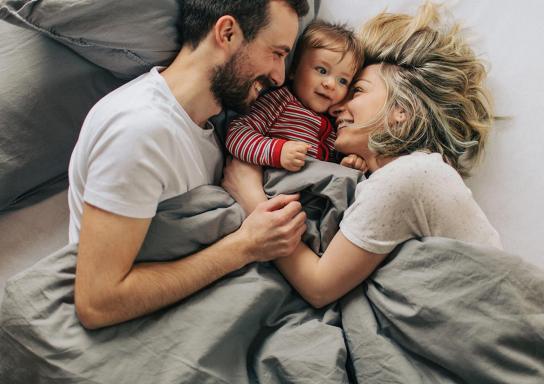 Paar liegt glücklich im Bett mit Kleinkind zwischen sich.