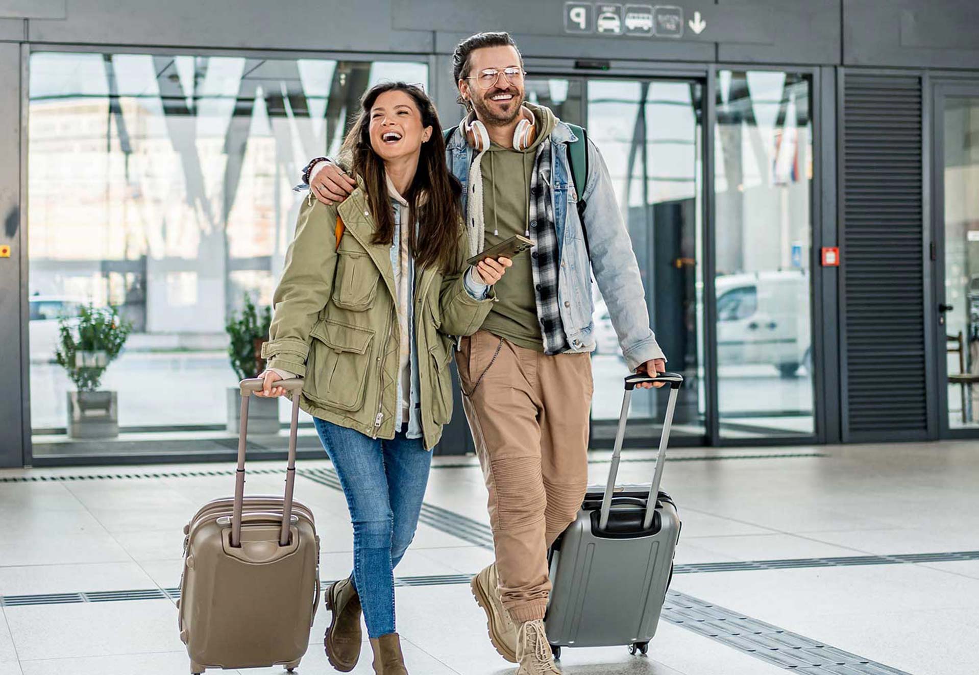 Ein Paar mit Rollkoffern und Smartphone steht vor einem modernen Gebäude, bereit für die Reise.