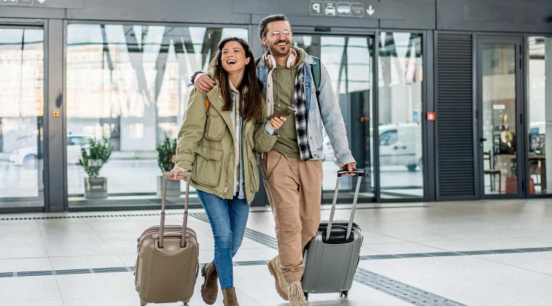 Ein Paar mit Rollkoffern und Smartphone steht vor einem modernen Gebäude, bereit für die Reise.