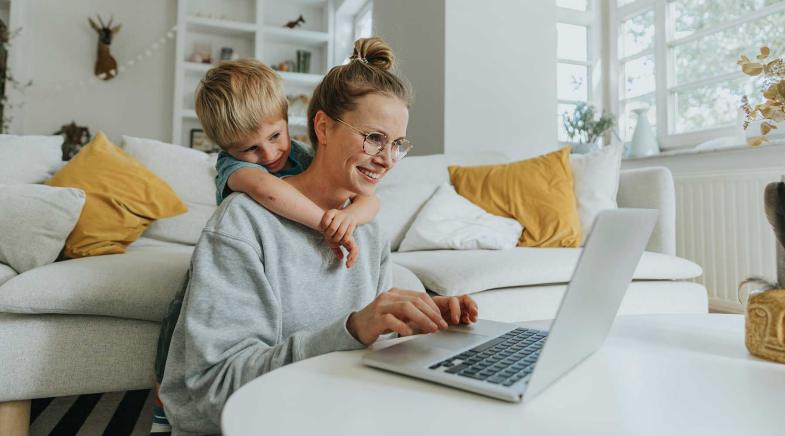 Frau sitzt vor ihrem Sofa und schaut auf ihren Laptop, ihr Sohn schaut ihr über die Schulter