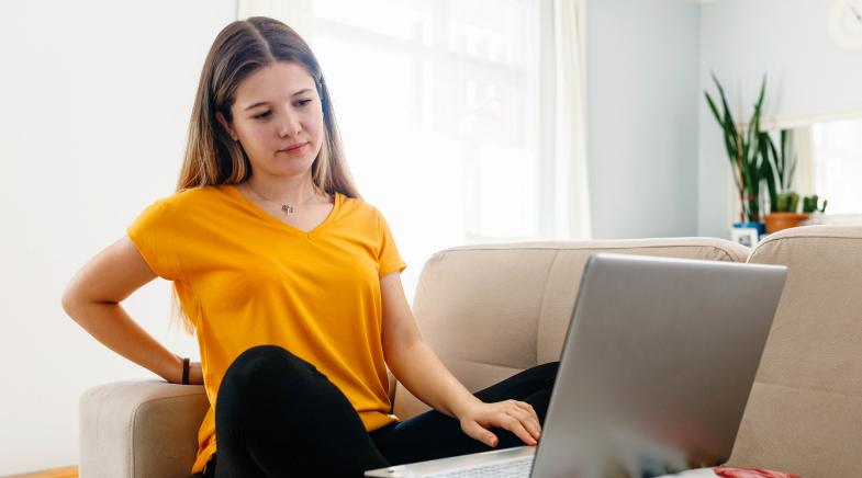 Junge Frau sitzt auf dem Sofa vor dem Laptop und hält sich den Rücken.