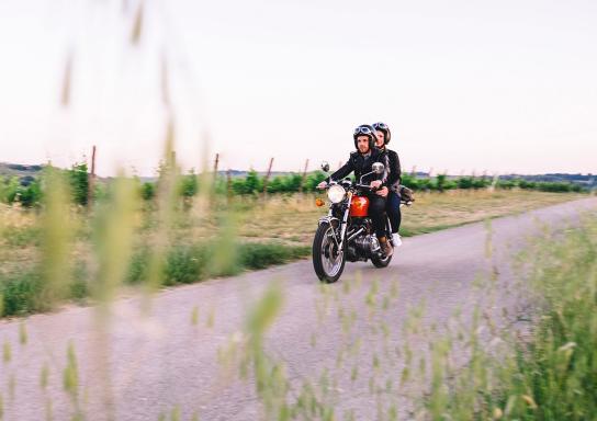 Ein Paar fährt mit einem alten Motorrad über einen Feldweg.