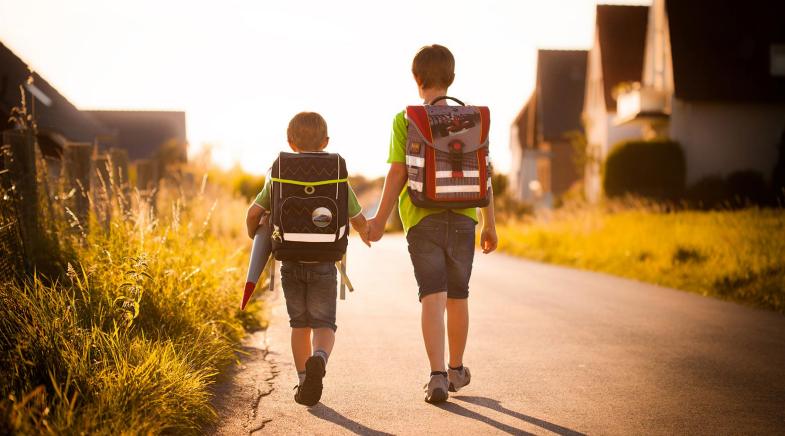 Zwei kleine Jungs mit Schulranzen gehen Hand in Hand durch ein Dorf.