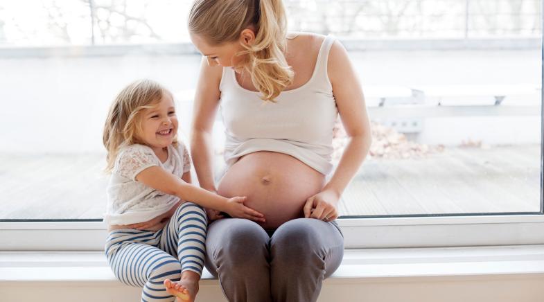 Kleines Mädchen fasst seiner Mutter an den schwangeren Bauch und lacht.