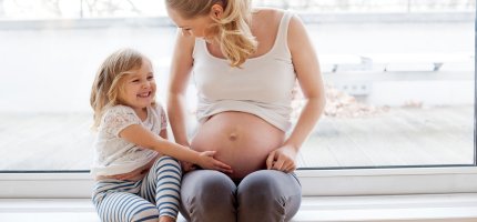 Kleines Mädchen fasst seiner Mutter an den schwangeren Bauch und lacht.