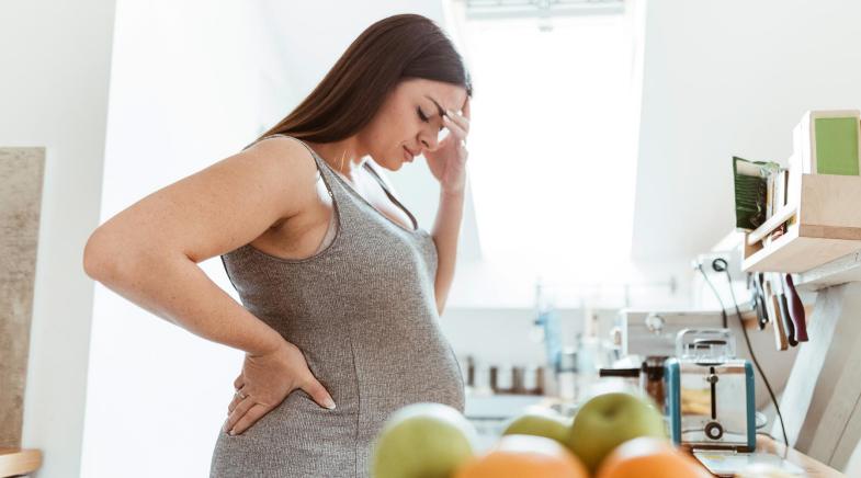 Eine schwangere Frau steht in der Küche und hält sich den Kopf.