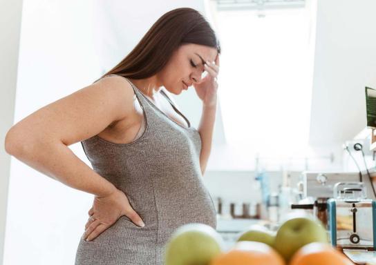 Eine schwangere Frau steht in der Küche und hält sich den Kopf.
