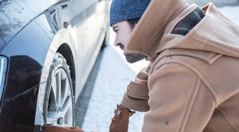 Eine Person trägt warme Kleidung und überprüft den Reifen eines Autos im Schnee.