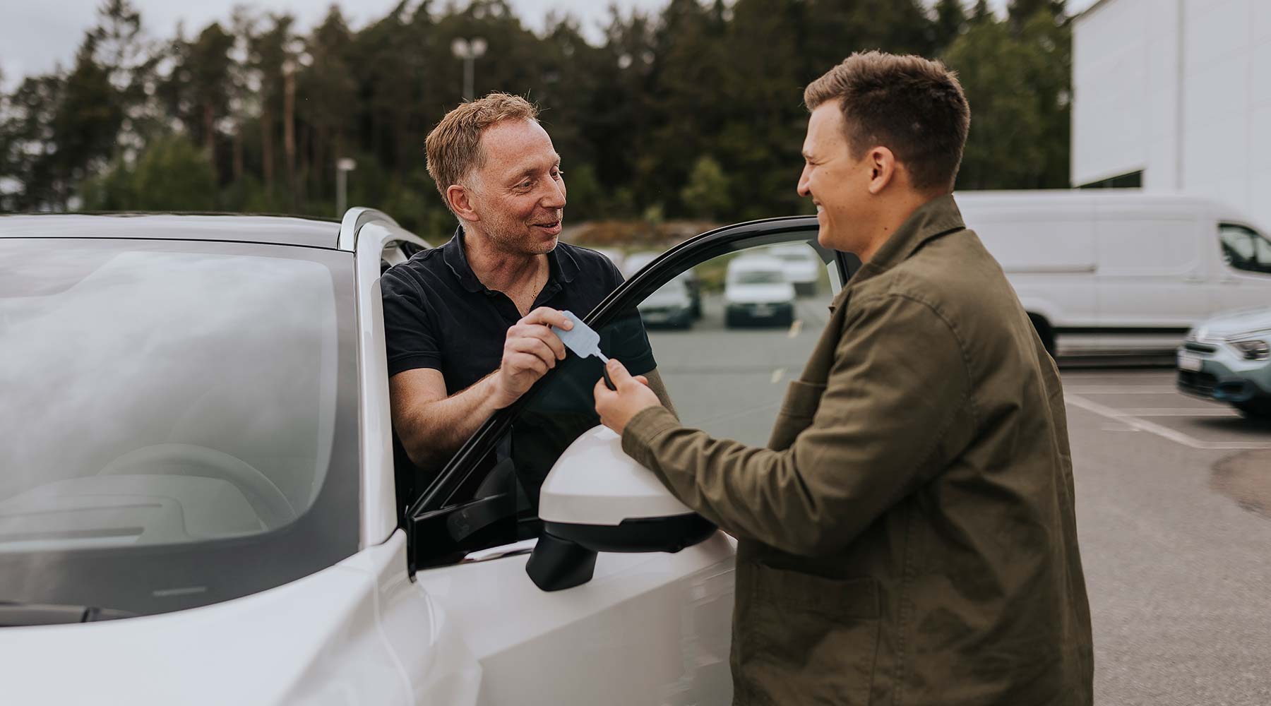 Zwei Personen stehen neben einem Auto auf einem Parkplatz. Eine Person übergibt der anderen eine Karte.