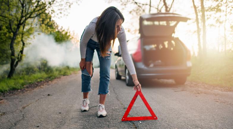 Eine Frau stellt auf einer Straße durch ein Waldstück ein Warndreieck hinter ihrem Auto auf.