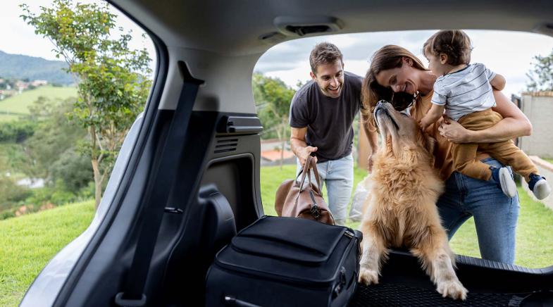 Ein Mann und eine Frau stehen mit einem Kleinkind und einem großen Hund vor einem geöffneten Kofferraum.