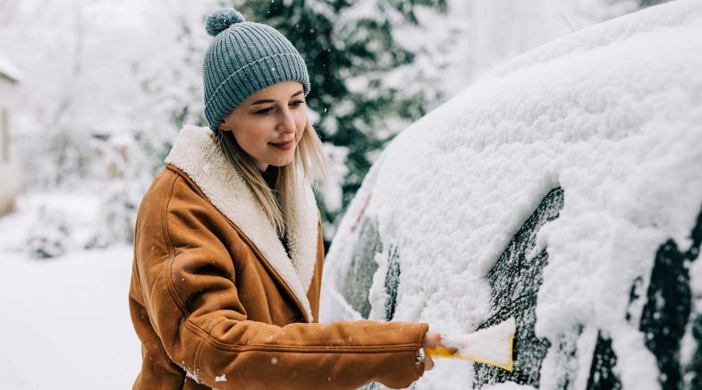 Eine Person entfernt Schnee von der Windschutzscheibe eines Autos in einer verschneiten Umgebung.