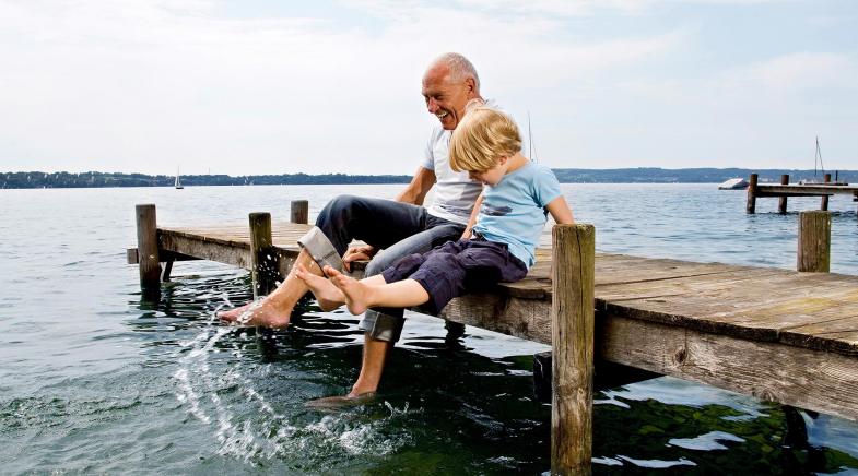 Großvater sitzt mit Enkel auf einem Steg am See und plantscht mit den Füßen im Wasser.