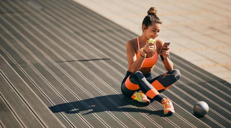 Eine Frau in Fitness-Kleidung sitzt entspannt auf dem Boden, isst einen Apfel und schaut aufs Handy.