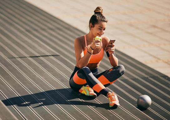 Eine Frau in Fitness-Kleidung sitzt entspannt auf dem Boden, isst einen Apfel und schaut aufs Handy.