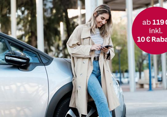 Eine Frau im Trenchcoat lehnt an ihrem Auto und schaut auf ihr Handy - daneben ein Störer mit der Aufschrift: ab 119 € inkl. 10 € Rabatt.