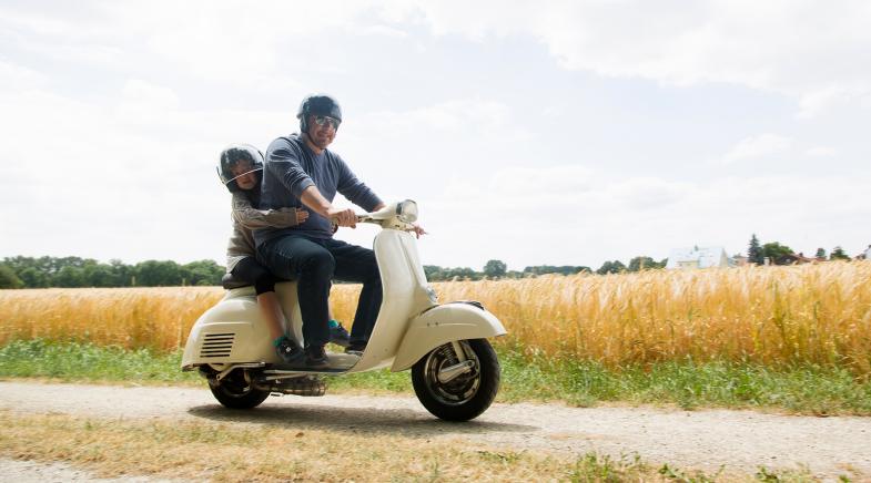 Ein Mann fährt mit einem Motorroller an einem Weizenfeld vorbei. Sein kleiner Sohn umarmt ihn von hinten.