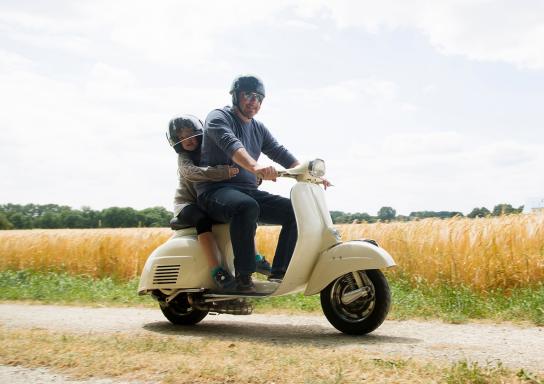 Ein Mann fährt mit einem Motorroller an einem Weizenfeld vorbei. Sein kleiner Sohn umarmt ihn von hinten.