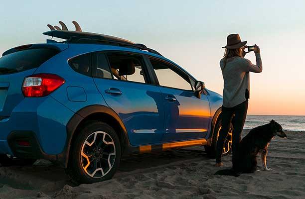 Eine Frau steht mit ihrem Hund und ihrem Auto am Strand und macht ein Foto vom Sonnenuntergang am Meer.