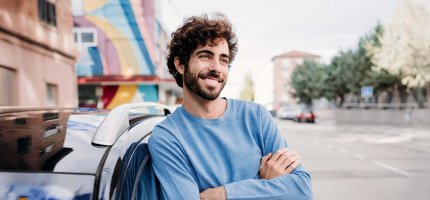 Lächelnder Mann mit verschränkten Armen, der an einem Auto gelehnt vor sich hin träumt.