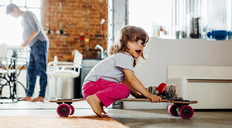 Kleines Mädchen sitzt in in der Wohnung auf einem Skateboard während seine Mutter nicht hinsieht.