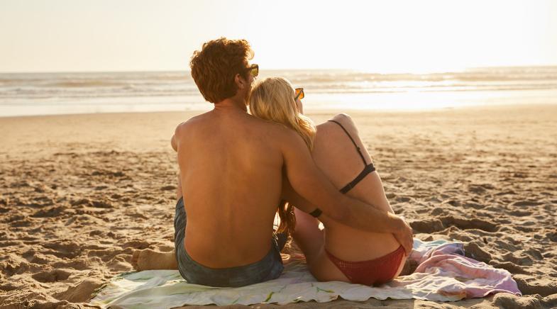 Mann in Badehose umarmt eine Frau im Bikini während beide am Strand sitzen und aufs Meer schauen.